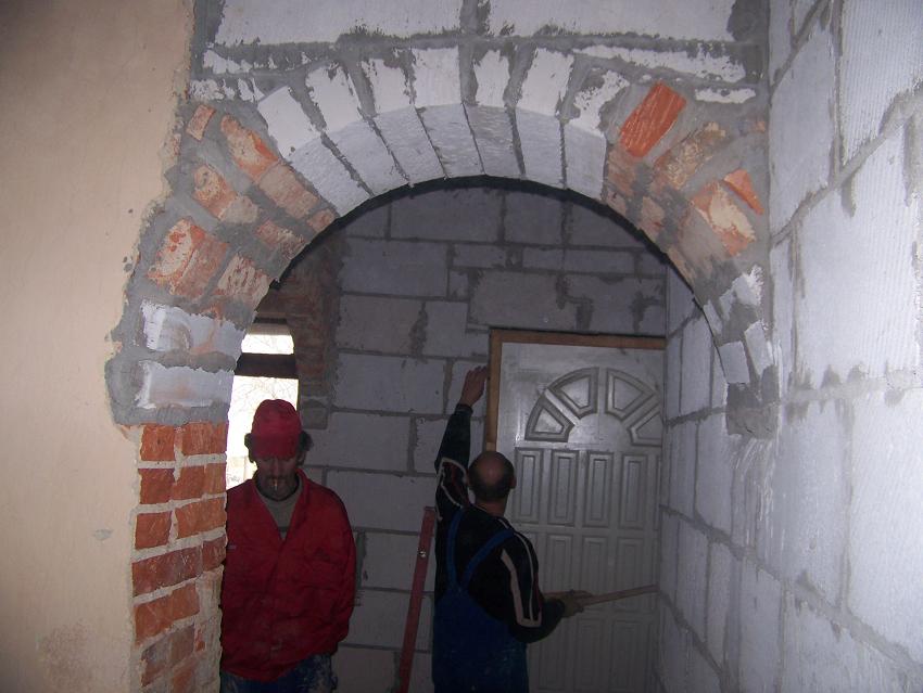 Wnętrze w trakcie budowy, widoczny murarz w czerwonej kurtce, łukowe przejście z cegieł i bloczków, ściany z białych bloczków, drzwi w tle, etap wznoszenia ścian działowych.